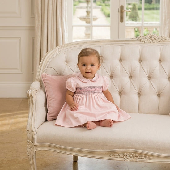Carriage Boutique baby girl wearing a pink taffeta dress with hand-smocked floral details and puffed sleeves on a sofa