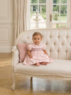 Carriage Boutique baby girl wearing a pink taffeta dress with hand-smocked floral details and puffed sleeves on a sofa