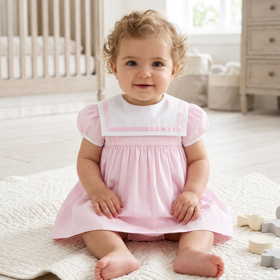 Carriage Boutique baby girl wearing a pink and white short sleeve dress with a classic sailor collar in a nursery.