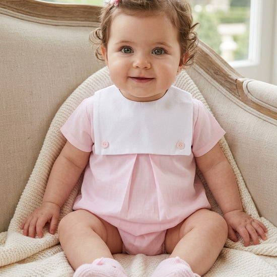 Carriage Boutique baby girl bubble romper in light pink and white cotton with a button bib, worn by a smiling baby.