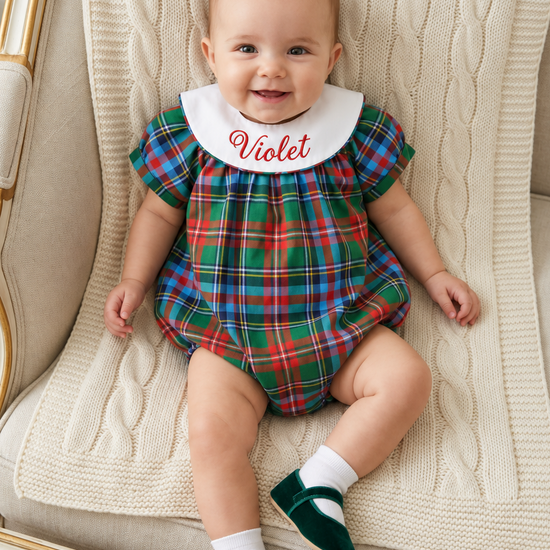 Carriage Boutique baby girl wearing a multicolor plaid bubble romper with a white monogrammed collar on a cream chair