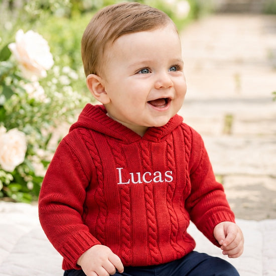 Carriage Boutique red cable knit hooded baby sweater with white monogrammed name on chest worn by a smiling toddler