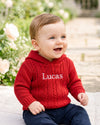 Carriage Boutique red cable knit hooded baby sweater with white monogrammed name on chest worn by a smiling toddler
