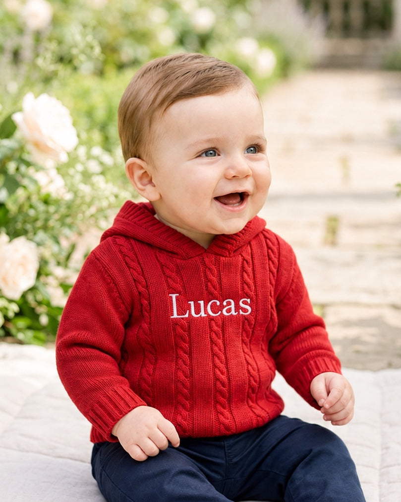 Carriage Boutique red cable knit hooded baby sweater with white monogrammed name on chest worn by a smiling toddler