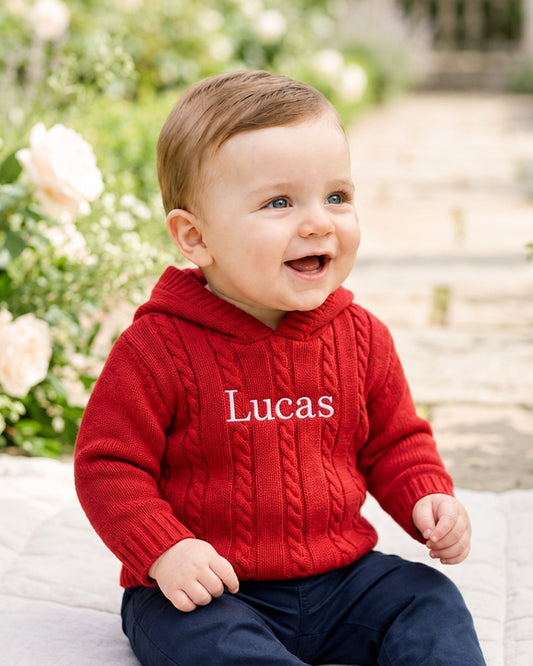 Carriage Boutique red cable knit hooded baby sweater with white monogrammed name on chest worn by a smiling toddler