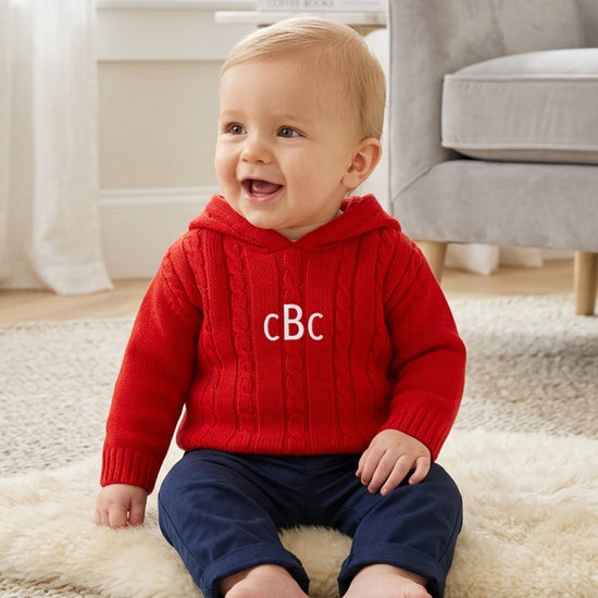 Carriage Boutique red cable knit hooded baby sweater with monogram on chest worn by a smiling baby on a rug
