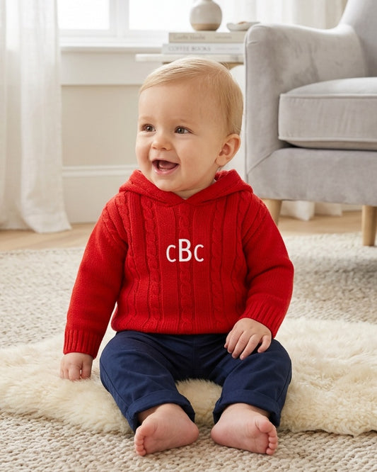 Carriage Boutique red cable knit hooded baby sweater with monogram on chest worn by a smiling baby on a rug