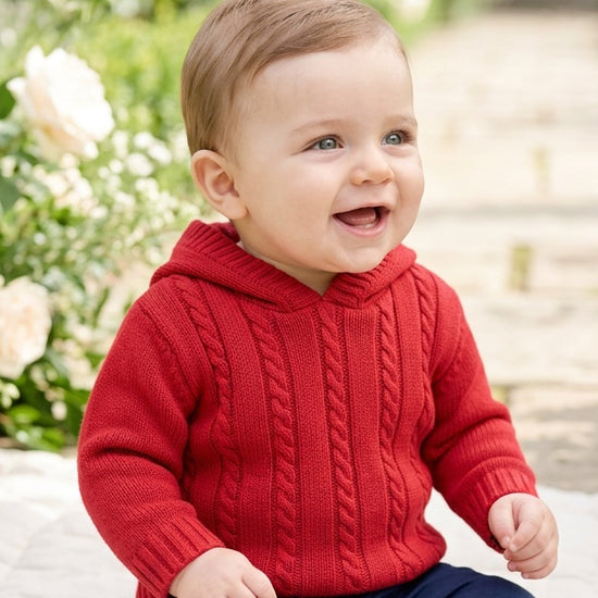 Carriage Boutique red cable knit hooded baby sweater worn by a smiling toddler in a garden setting
