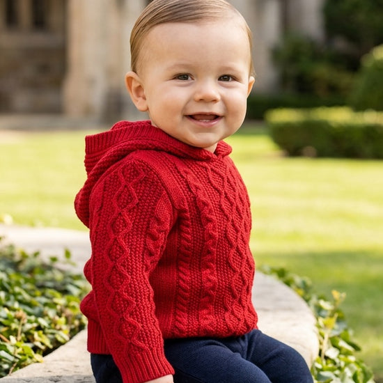 Carriage Boutique red cable knit hooded sweater worn by a smiling toddler sitting on a stone wall outdoors.