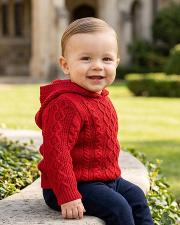 Carriage Boutique red cable knit hooded sweater worn by a smiling toddler sitting on a stone wall outdoors.