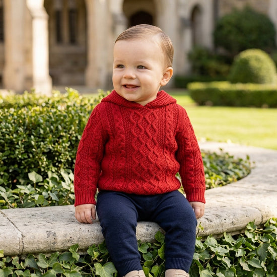 Carriage Boutique red cable knit hooded sweater worn by a smiling toddler sitting on a stone wall outdoors.