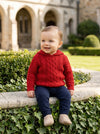 Carriage Boutique red cable knit hooded sweater worn by a smiling toddler sitting on a stone wall outdoors.
