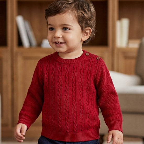 Toddler wearing a Carriage Boutique red crew neck cable knit sweater with shoulder buttons in a cozy room.