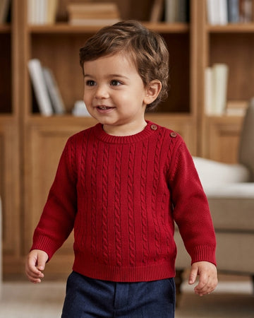 Toddler wearing a Carriage Boutique red crew neck cable knit sweater with shoulder buttons in a cozy room.