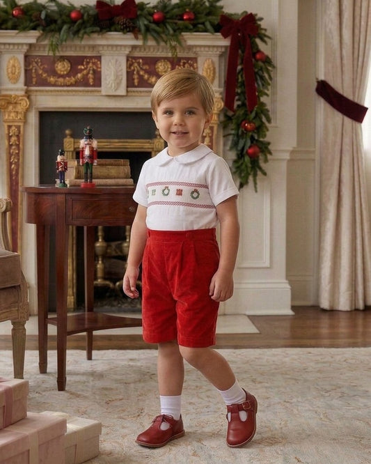 Carriage Boutique boy wearing a white smocked shirt with Christmas motifs and red corduroy shorts in a festive room.