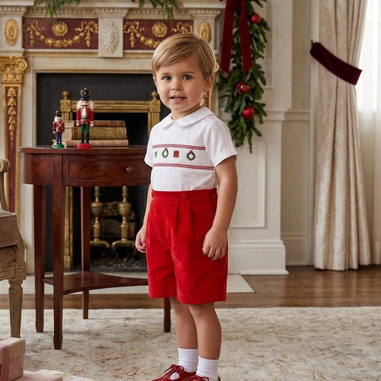Carriage Boutique boy wearing a white smocked shirt with Christmas motifs and red corduroy shorts in a festive room.