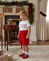 Carriage Boutique boy wearing a white smocked shirt with Christmas motifs and red corduroy shorts in a festive room.