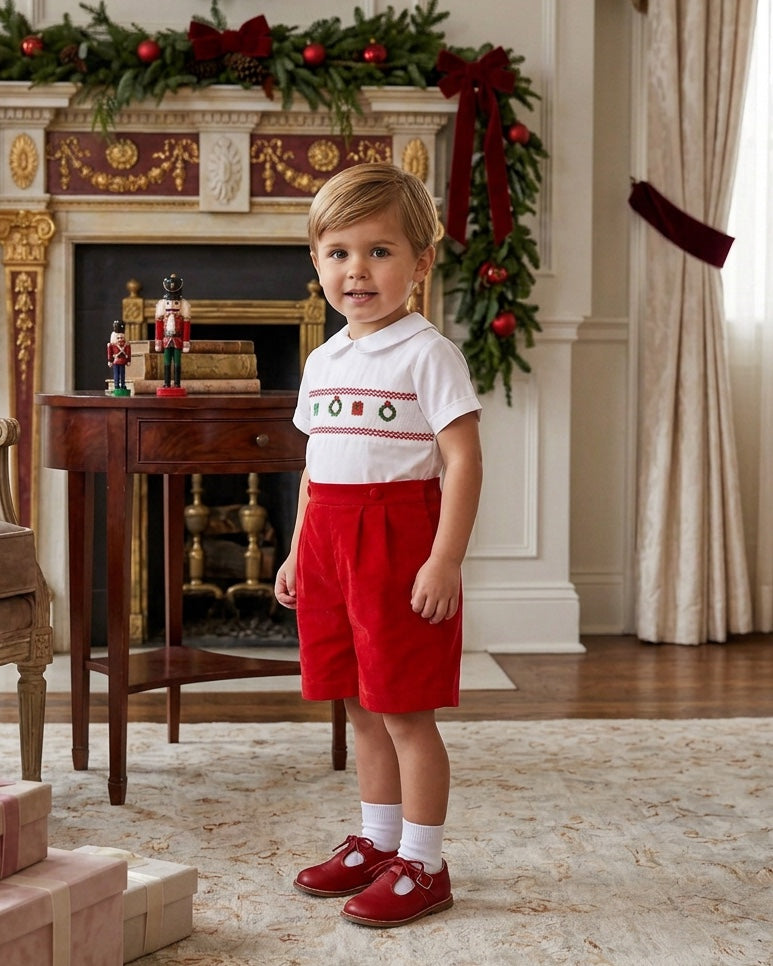 Carriage Boutique boy wearing a white smocked shirt with Christmas motifs and red corduroy shorts in a festive room.