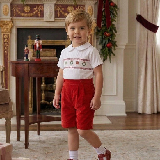 Carriage Boutique boy wearing a white smocked shirt with Christmas motifs and red corduroy shorts in a festive room.