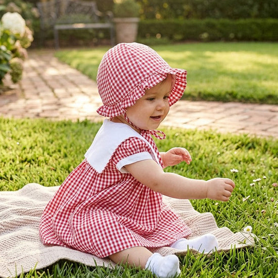 Carriage Boutique baby girl wearing a red gingham sun hat with a matching dress, sitting on a blanket in a garden.