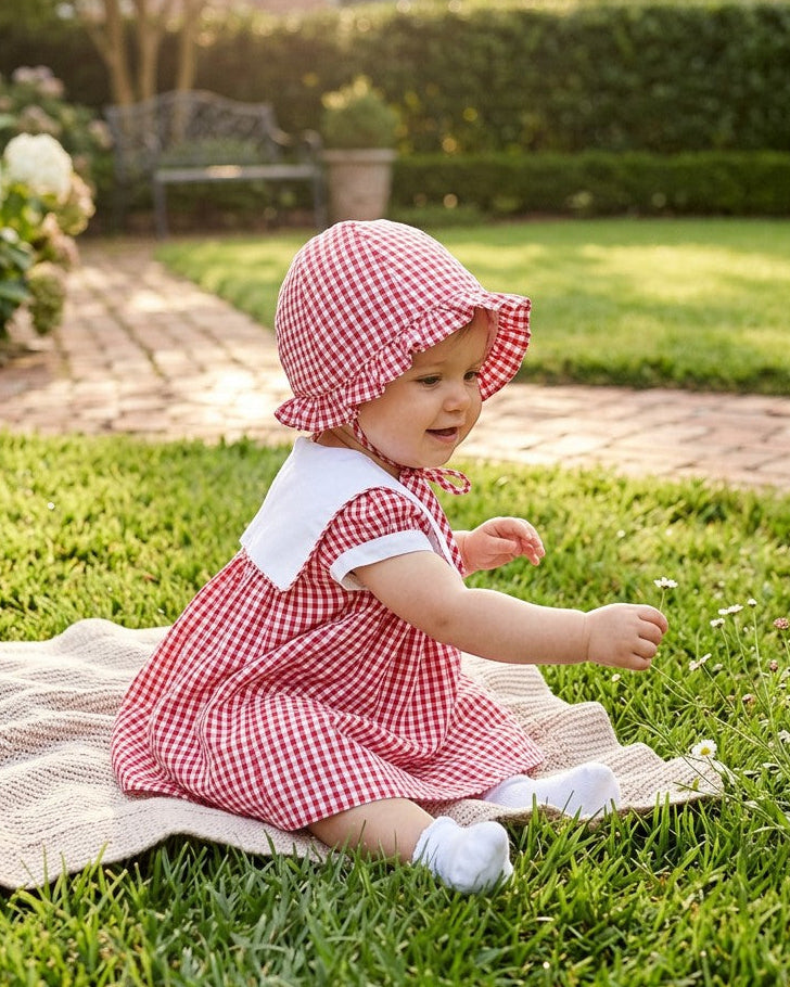 Carriage Boutique baby girl wearing a red gingham sun hat with a matching dress, sitting on a blanket in a garden.