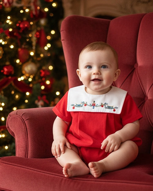 Baby boy wearing a Carriage Boutique red cotton bubble romper with a smocked nutcracker bib sitting in a red chair.