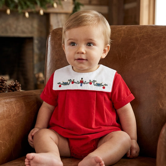 Carriage Boutique red cotton baby boy bubble romper with smocked nutcracker bib sitting in a leather chair