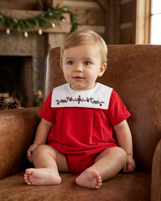 Carriage Boutique red cotton baby boy bubble romper with smocked nutcracker bib sitting in a leather chair