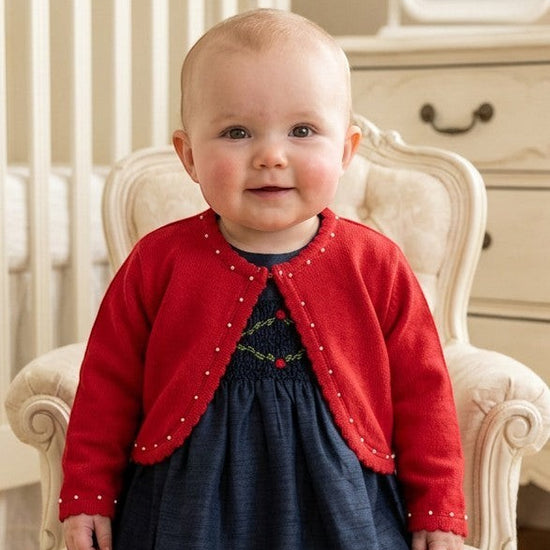 Carriage Boutique baby girl red bolero sweater with pearl trim worn over a navy dress in a nursery setting