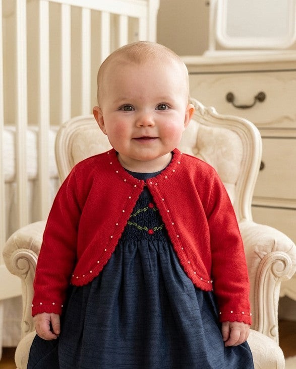 Carriage Boutique baby girl red bolero sweater with pearl trim worn over a navy dress in a nursery setting