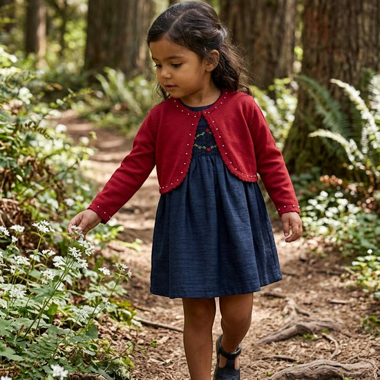 Carriage Boutique toddler girl wearing a red bolero sweater with pearl accents over a navy dress in a forest.