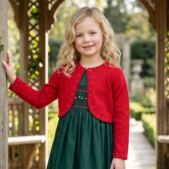 Carriage Boutique red bolero sweater with pearl accents worn by a young girl over a green dress in a garden.