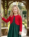 Carriage Boutique red bolero sweater with pearl accents worn by a young girl over a green dress in a garden.