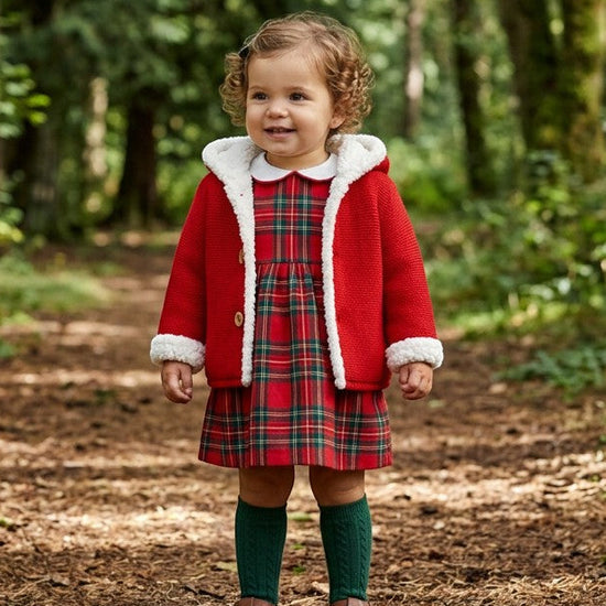 Toddler wearing a Carriage Boutique red knit hooded jacket with white sherpa lining over a plaid dress in a forest