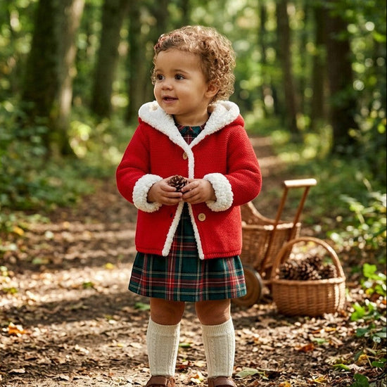 Toddler wearing a red Carriage Boutique sherpa-lined hooded knit jacket with a plaid dress in a forest setting