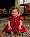Carriage Boutique red silk baby dress with smocked floral embroidery and puffy sleeves, worn by a toddler on a rug.