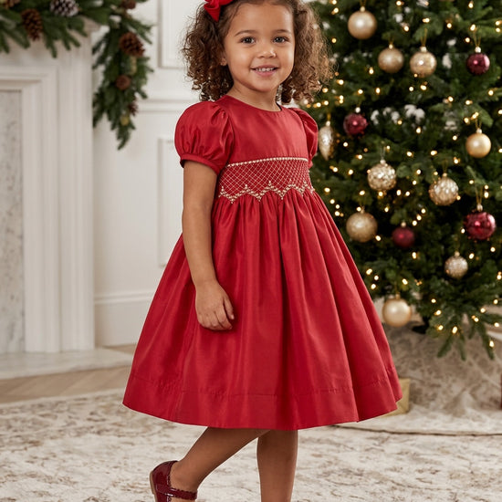 Carriage Boutique toddler girl in a red taffeta dress with smocked diamond detailing and puffed sleeves by a tree