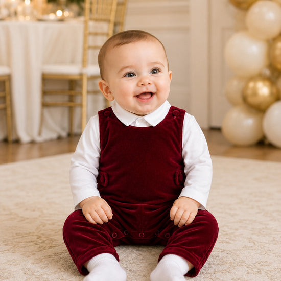 Carriage Boutique baby boy wearing a red velvet long romper over a white shirt, sitting on a rug at a party.