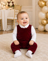 Carriage Boutique baby boy wearing a red velvet long romper over a white shirt, sitting on a rug at a party.