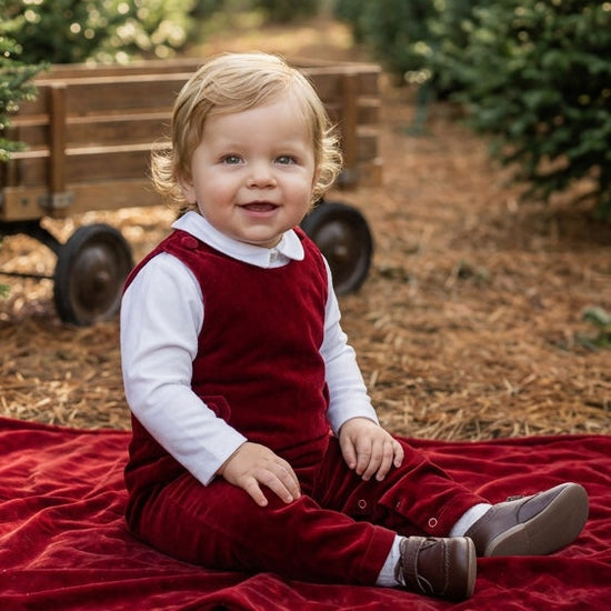Carriage Boutique baby boy wearing a red velvet long romper over a white shirt, sitting on a red blanket outdoors.