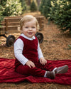 Carriage Boutique baby boy wearing a red velvet long romper over a white shirt, sitting on a red blanket outdoors.