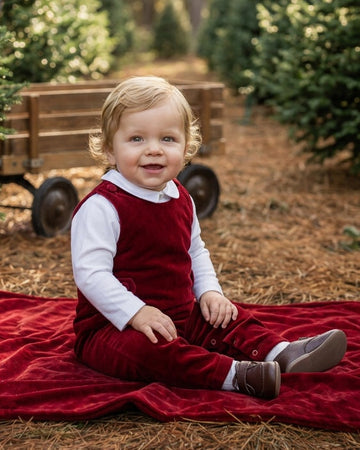 Carriage Boutique baby boy wearing a red velvet long romper over a white shirt, sitting on a red blanket outdoors.