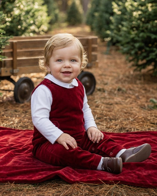 Carriage Boutique baby boy wearing a red velvet long romper over a white shirt, sitting on a red blanket outdoors.
