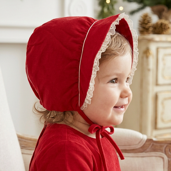 Carriage Boutique red velvet baby bonnet with white lace trim and tie closure worn by a smiling toddler