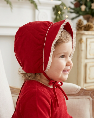Carriage Boutique red velvet baby bonnet with white lace trim and tie closure worn by a smiling toddler