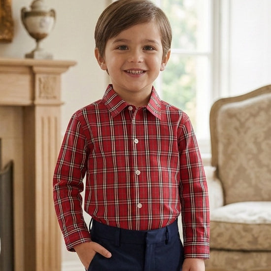 A young boy smiling while wearing a Carriage Boutique red and white plaid long sleeve cotton shirt with navy pants.
