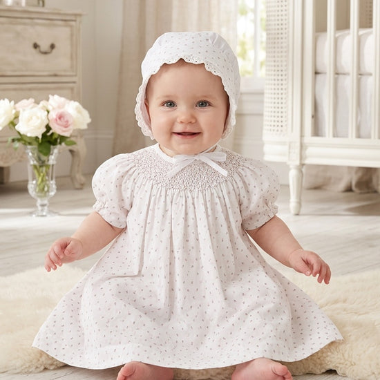 Carriage Boutique baby girl wearing a white dress with a delicate rosebud print and matching bonnet in a nursery.