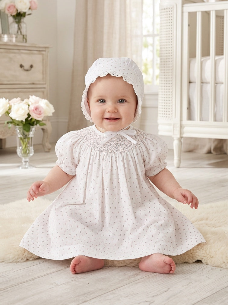 Carriage Boutique baby girl wearing a white dress with a delicate rosebud print and matching bonnet in a nursery.