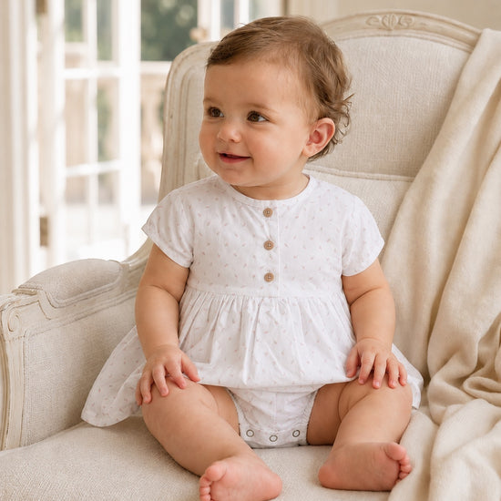 Carriage Boutique white baby girl dress with delicate rosebud print, cap sleeves, and wooden buttons on a seated baby.