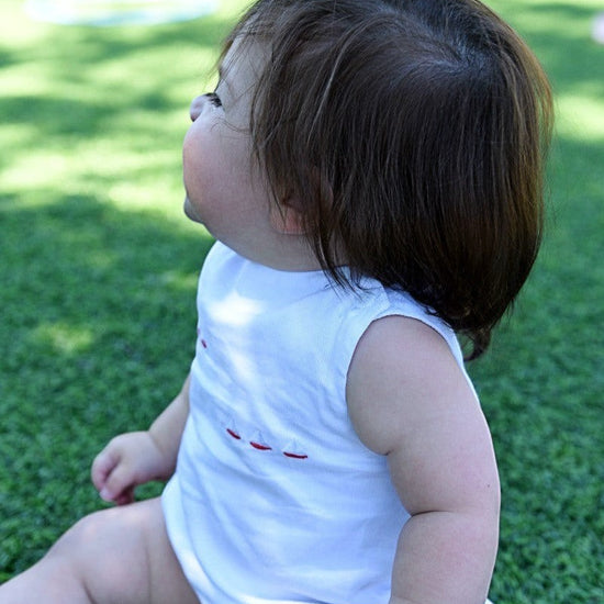 Carriage Boutique white sleeveless bubble with smocked detailing and embroidered red sailboats. Baby sitting on grass.